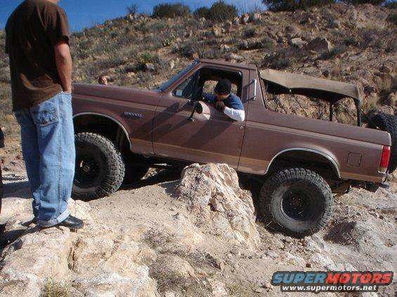 Brown Ford Bronco with a tan soft top is climbing over rocks on a trail. Driver is visible. Another person stands to the left