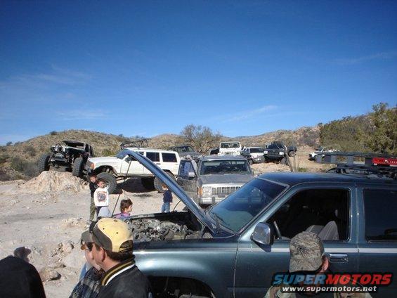 Dark green SUV with hood open, parked on dirt. Multiple off-road vehicles in background