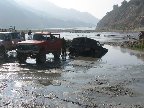 Red pickup truck with lifted suspension and off-road tires, parked next to a black SUV partially submerged in water and mud