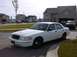 1999 Ford Crown Victoria - A white sedan is parked in front of a suburban house with a garage containing another vehicle.