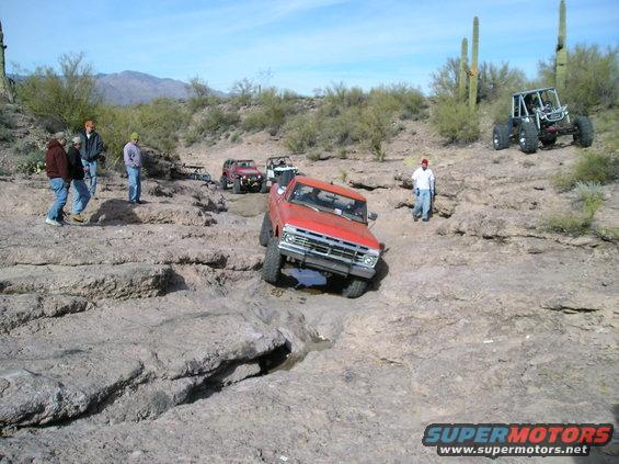 Red pickup truck and other off-road vehicles on a rocky trail with cacti and mountains in the background