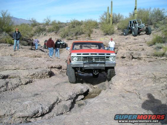 Red Ford pickup truck navigating rocky terrain with other off-road vehicles and people around