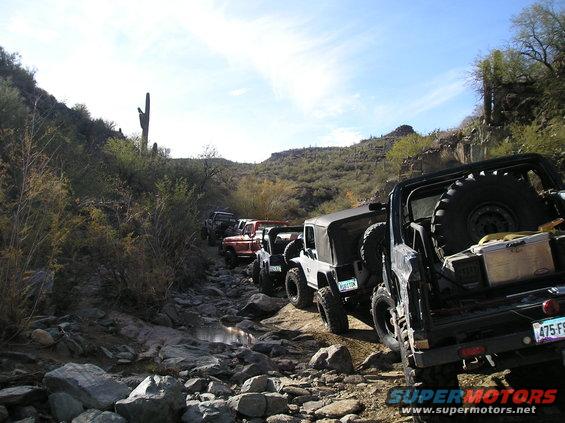 Line of off-road vehicles navigating a rocky, dry creek bed with desert vegetation and mountains in the background