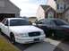 1999 Ford Crown Victoria - Two Ford Crown Victorias, one white and one dark colored, are parked in a driveway in front of houses.