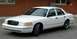 1999 Ford Crown Victoria - A white sedan is parked outside a brick building. The front of the car is angled towards the camera.