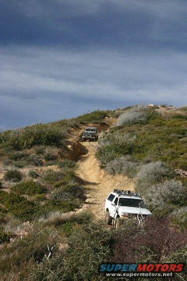 White SUV with roof rack climbing a dirt trail, another vehicle behind it