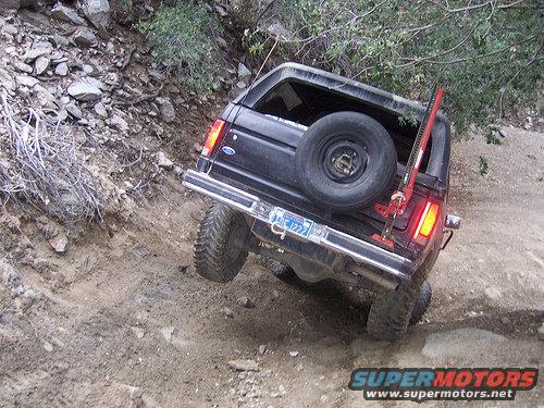 Black Ford Bronco on a dirt trail, tilted to the side. A Hi-Lift jack is mounted vertically on the rear