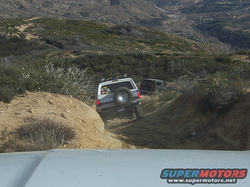 Silver 1981 Ford Bronco driving on a dirt trail, rear view. Another SUV follows