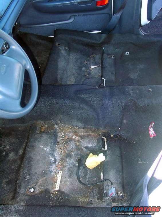 Car interior floor with visible rust and debris, showing part of the steering wheel and door panel