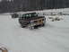 1979 Ford Bronco - Two SUVs navigate a snowy path with trees and a wooden fence in the background.