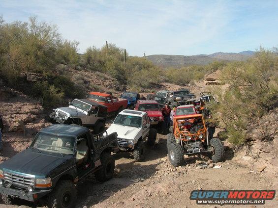 Dark green pickup truck in foreground, followed by a line of off-road vehicles on a dirt path
