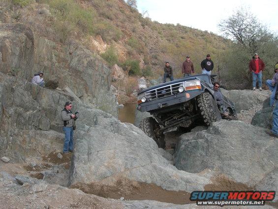 Off-road truck with large tires climbing over rocks, people watching