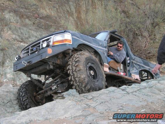 Modified 1992 Ford F-150 on rocky terrain, driver visible. Aftermarket bumper and large tires