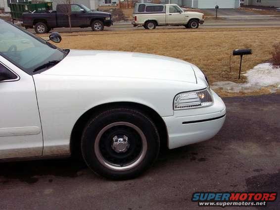 White sedan front quarter panel and wheel, with a dark pickup truck and a white pickup truck with camper shell in the background