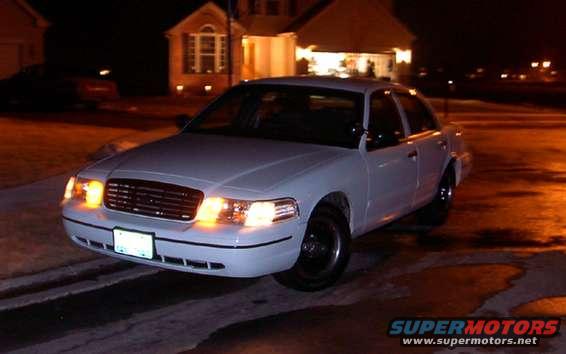 White sedan, front 3/4 view, parked at night. Headlights on, license plate visible