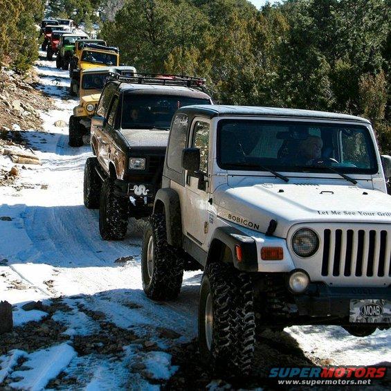 White Jeep Wrangler with 'BOBICON' and 'Let Me See Your Grill' text, followed by other off-road vehicles on a snowy mountain road