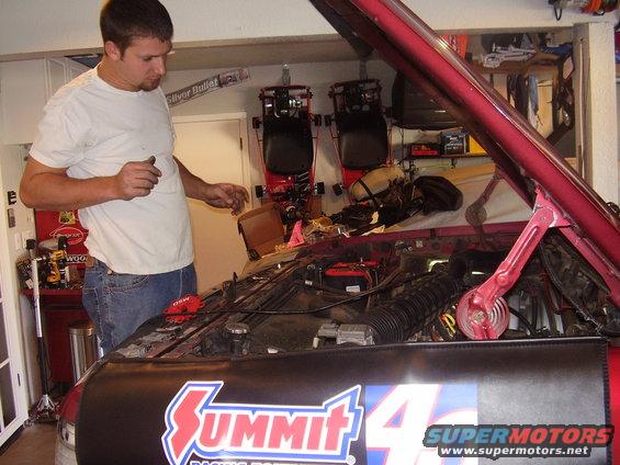 Man in white t-shirt and jeans working on red car engine in garage. Summit Racing logo on fender cover