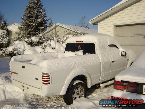 White pickup truck covered in snow, parked next to a garage. Another car is partially visible on the right
