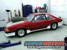 Red and white 1990 Ford Mustang with a large hood scoop, chrome wheels, and white body kit, viewed from the front-right in a garage