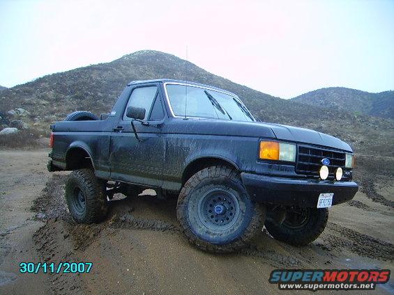 1990 Ford Bronco driving through mud with hills in background