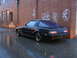 1983 Mazda RX-7 - A black 1983 Mazda RX-7 sits parked on a wet, reflective street beside a graffiti-covered brick wall.