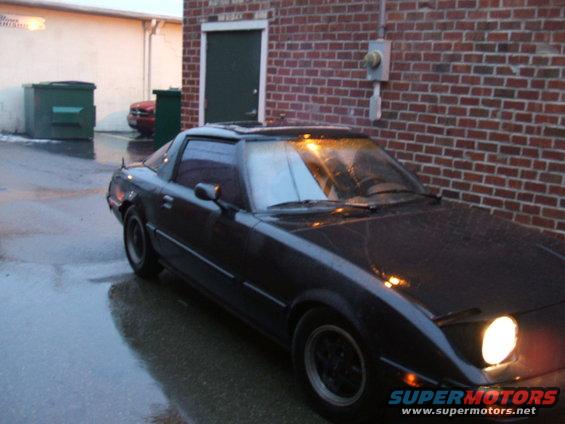1983 Mazda RX-7 parked on wet asphalt near a brick building, front passenger side visible with headlight on