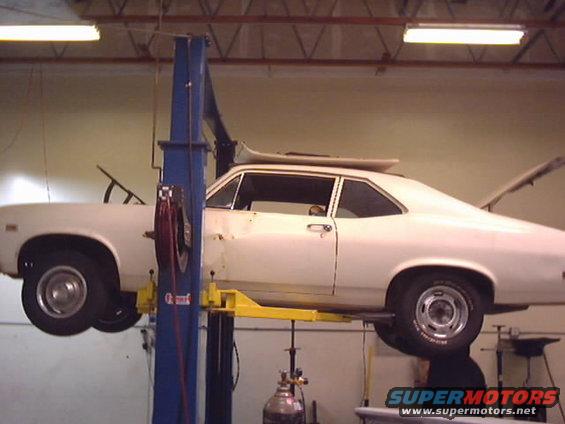 White coupe on a car lift, rear trunk open. Appears to be a 1970s model