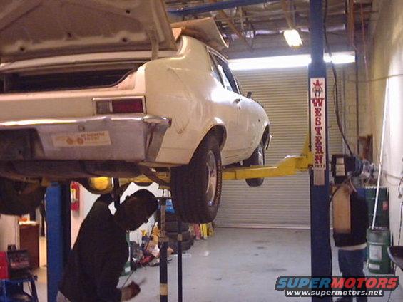 White sedan on a car lift, rear view, showing trunk open and a sticker on the bumper