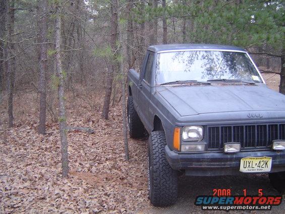 1988 Jeep Comanche pickup truck parked on a leaf-covered ground in a wooded area. Front grille and headlights visible