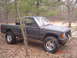 1988 Jeep Comanche - A 1988 Jeep Comanche pickup truck sits in a forest clearing, its dark gray paint contrasting with the natural surroundings.