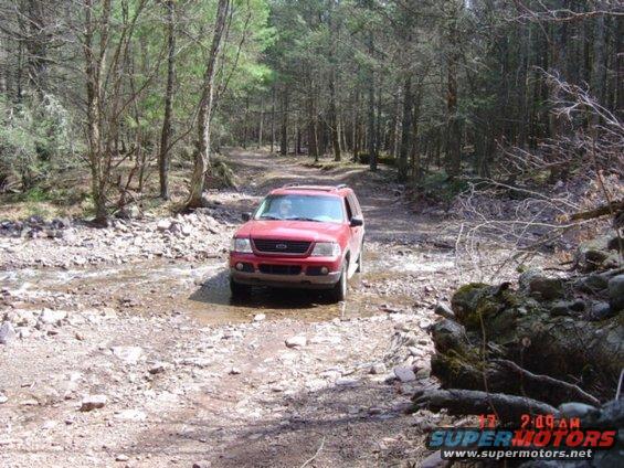 Red 2002 Ford Explorer driving through shallow water on a dirt road in a forest