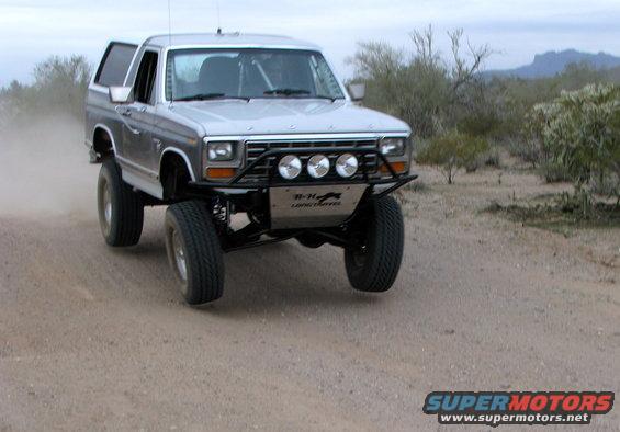 Silver 1980 Ford Bronco with off-road modifications driving on a dirt road, kicking up dust