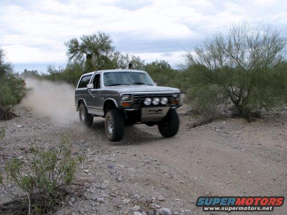 1980 Ford Bronco driving on a dusty desert road with aftermarket lights and bumper