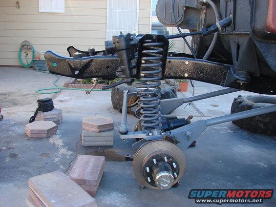 Vehicle front suspension assembly with coil spring and brake rotor visible. Frame section is black