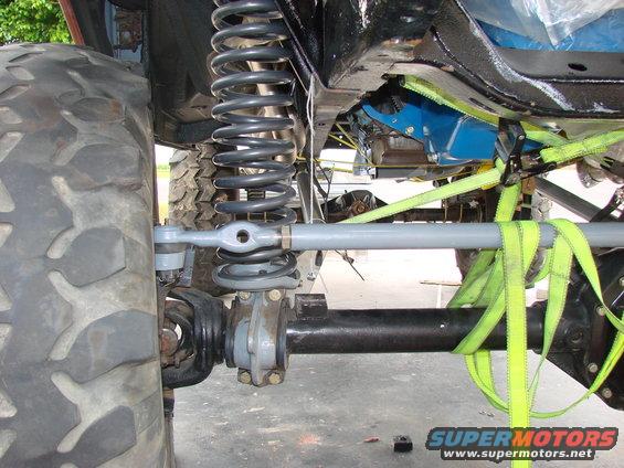 Close-up of a vehicle's front suspension with large tire, coil spring, and steering linkage, secured by yellow straps