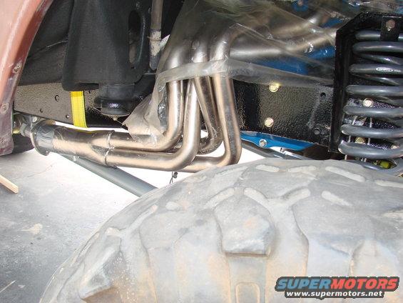 Close-up of shiny exhaust headers and suspension parts under a vehicle, with a large tire in the foreground