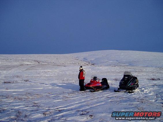 Red and black snowmobiles parked in a vast, snow-covered field under a clear blue sky