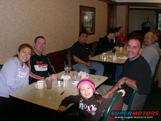 Group of people, including a child in a pink hat, seated at tables in a restaurant. A man in a black shirt smiles at the camera