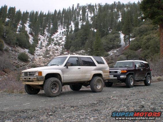 White 1980s Toyota 4Runner and a dark Toyota FJ Cruiser on a dirt road with snowy hills in the background