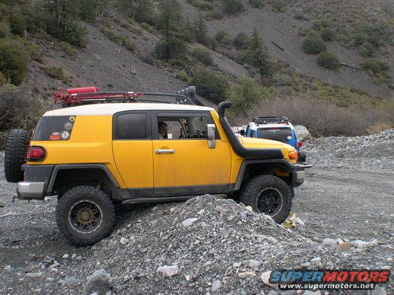 Yellow 2007 Toyota FJ Cruiser with snorkel and roof rack driving on a rocky trail. Another blue SUV is behind it