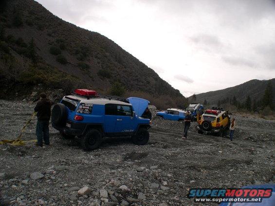 Several Toyota FJ Cruisers parked in a rocky, outdoor area with hills in the background. One has its hood up