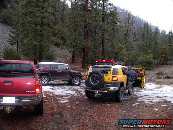 Yellow Toyota FJ Cruiser with spare tire on rear, parked in snowy woods next to a red pickup truck