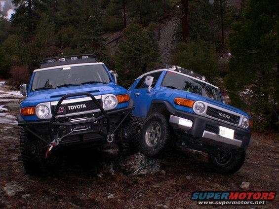 Two blue 2007 Toyota FJ Cruisers are driving on a rocky, wooded trail. One is in the foreground, the other is slightly behind and to the right