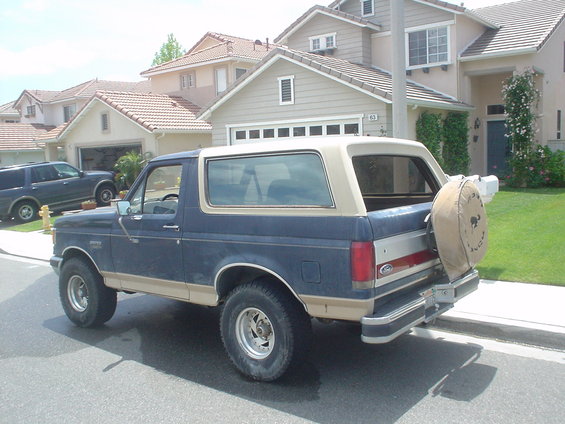 Blue and tan Ford Bronco parked on street, houses in background. Another SUV is parked in driveway