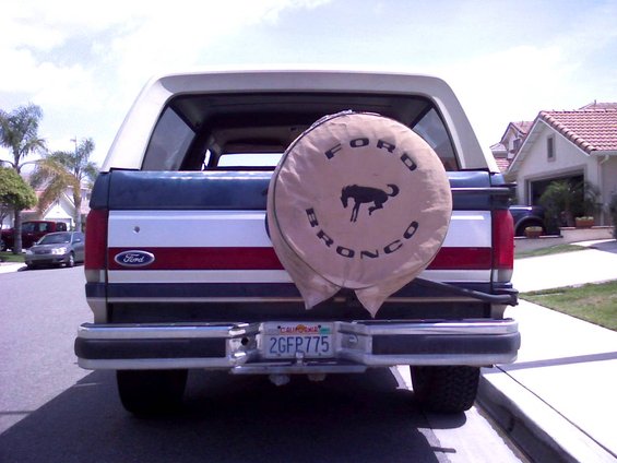 Rear of a blue and white Ford Bronco with a tan spare tire cover showing a horse logo and 'FORD BRONCO' text