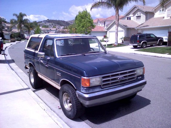 Dark blue Ford Bronco parked on a suburban street, with a dark SUV in a driveway