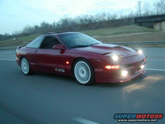 Red 1994 Ford Probe GTS driving on a highway with headlights on