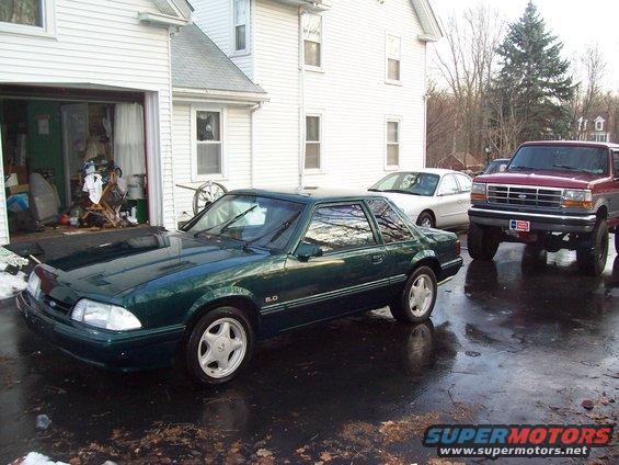 Green 1991 Ford Mustang coupe parked in driveway with a red Ford truck and white sedan behind it