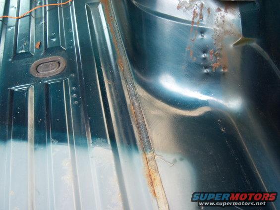 Interior floor of a vehicle showing ribbed metal panels and a rubber grommet. Some rust is visible
