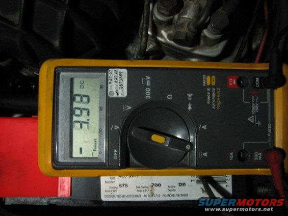 Yellow digital multimeter displaying -3.98 DC voltage, resting on a red object next to a car battery
